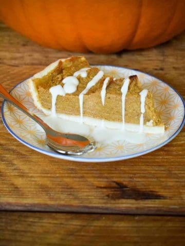 A piece of pumpkin pie on a small plate with a spoon next to it, oat cream drizzled over the pie