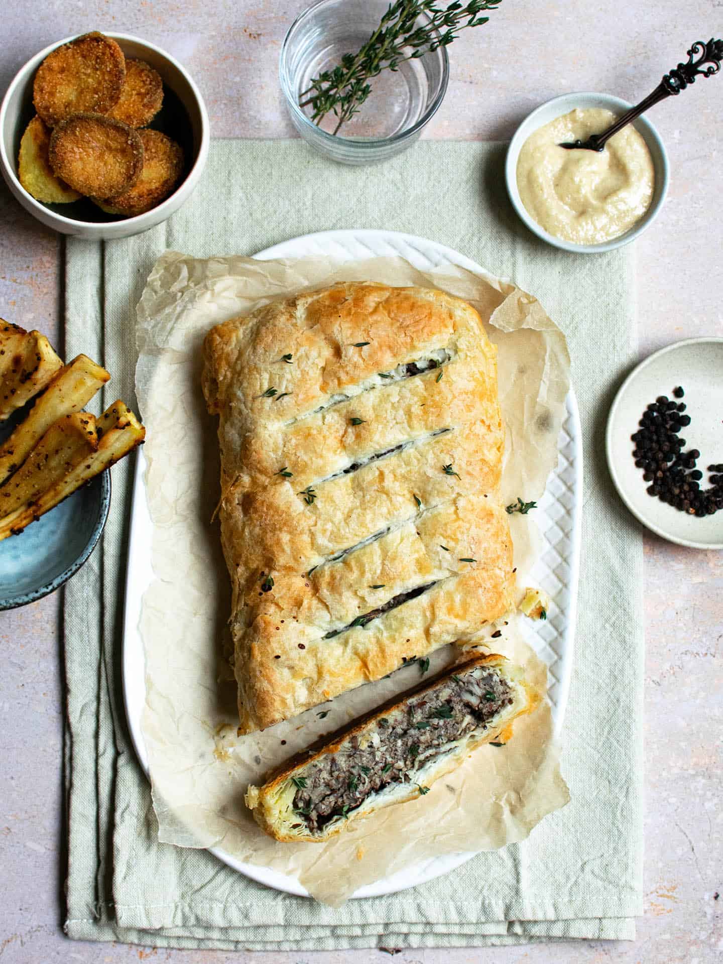 Vegan Mushroom Wellington on a white platter sitting on top of parchment paper, surrounded with vegan roast dinner dishes: parsnips, potatoes, sauce and peppercorns