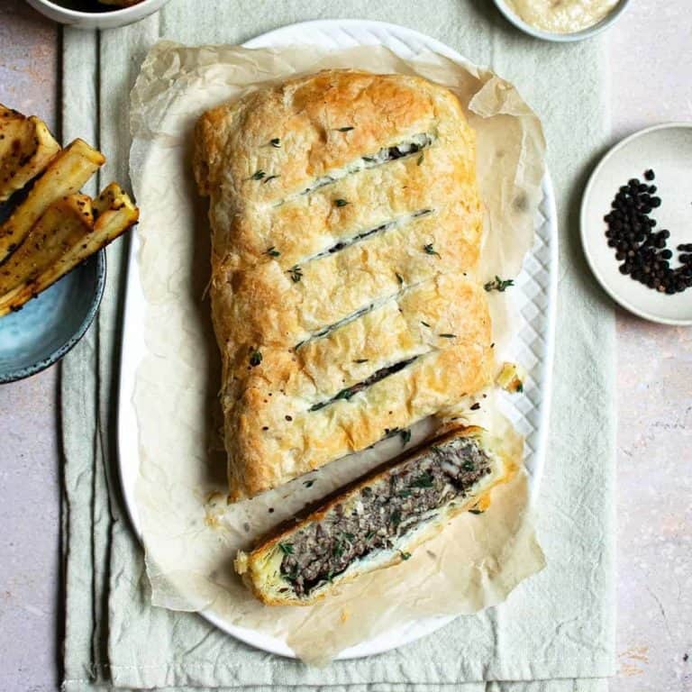Vegan Mushroom Wellington on a white serving plate surrounded by peppercorns, sauce and parsnips