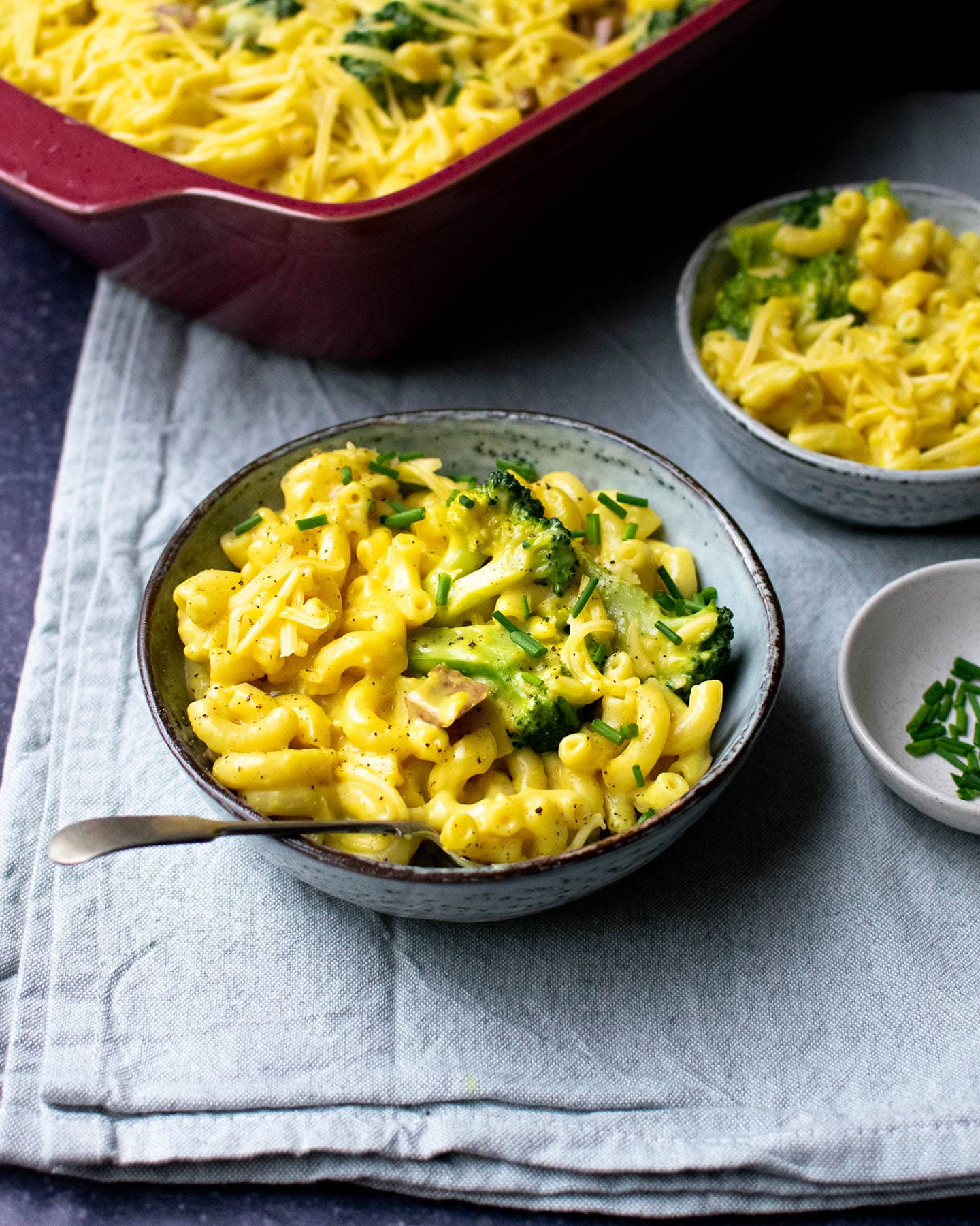 A blue bowl of baked vegan mac and cheese with broccoli in it, a small dish of chives next to it, a smaller bowl of food behind it and then a large red dish full of mac and cheese in the background