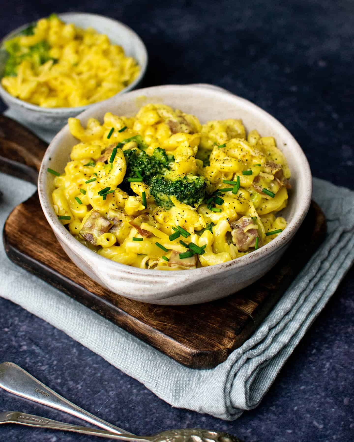 A bowl of nut free vegan mac and cheese with broccoli on top and some vegan bacon bits, a second smaller bowl is in the background