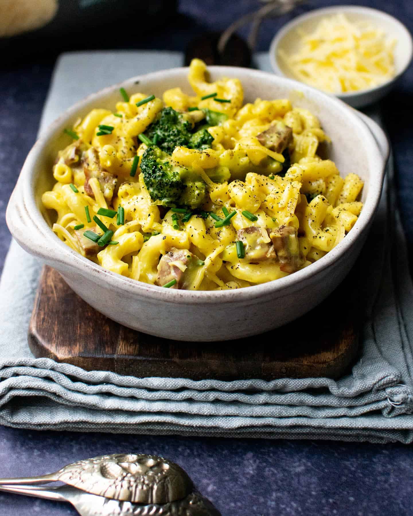 Baked vegan mac and cheese in a bowl sat on top of a chopping board with vegan cheese in the background and spoons in the foreground