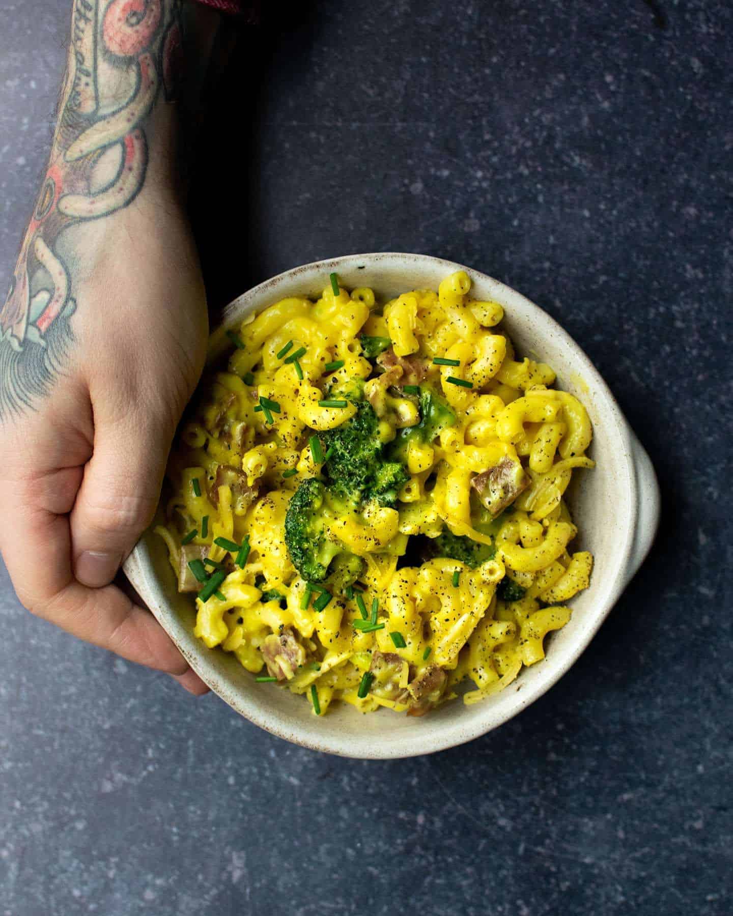A hand holding a bowl of baked vegan mac and cheese with broccoli and vegan bacon