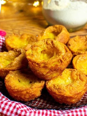 A pile of vegan Yorkshire puddings on a gingham cloth. In the background is a jar of chickpea or gram flour and some mechanical kitchen scales