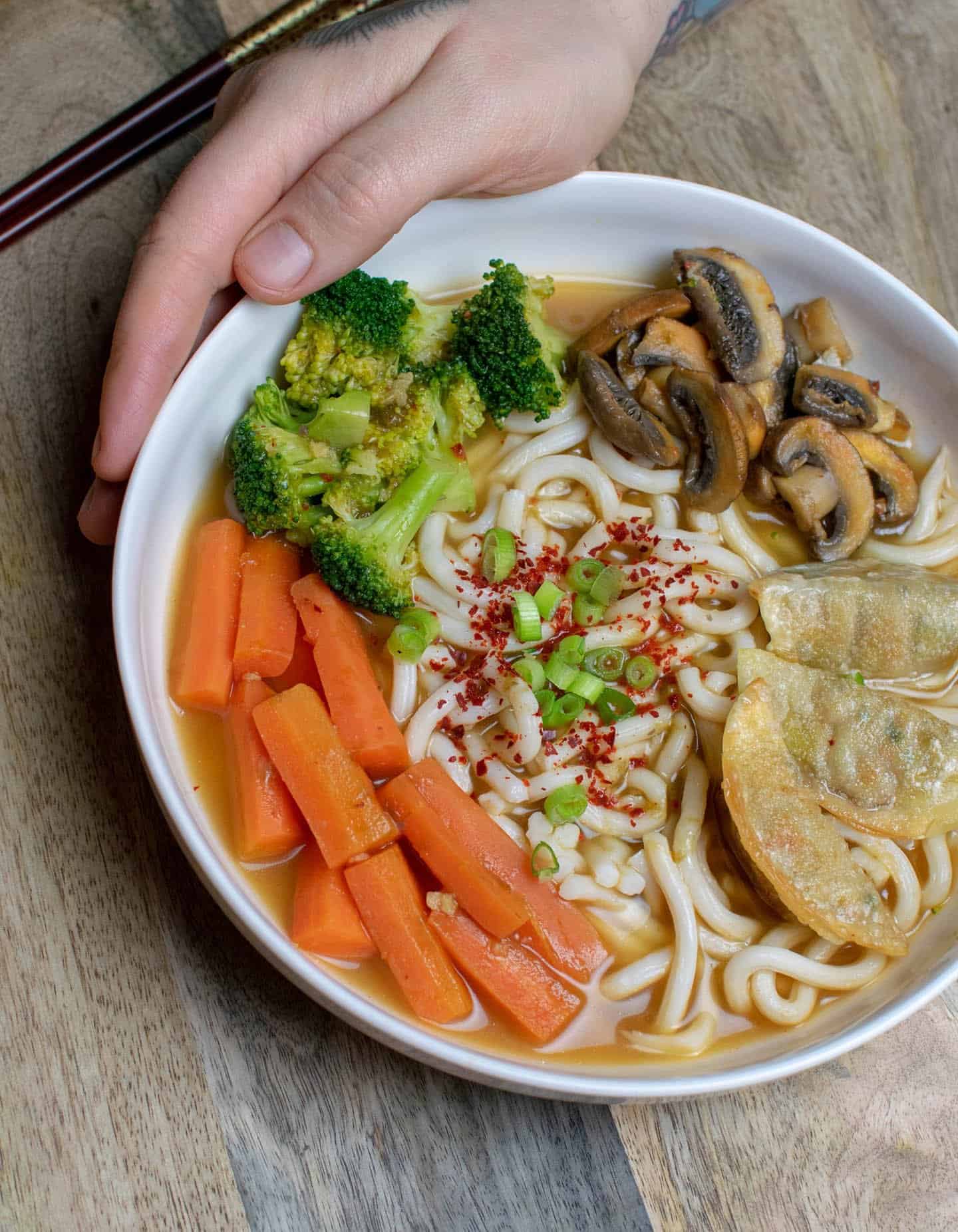 A white bowl full with ramen, broth, mushrooms, broccoli, carrots and gyozas is being cradled by a hand.