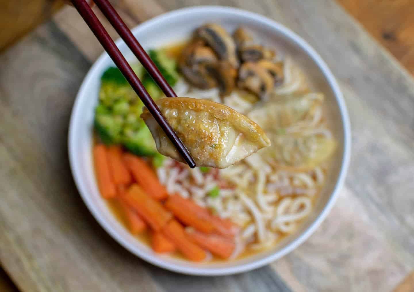 A bowl of ramen is blurred out in the background and in the foreground a hand is holding a pair of chopsticks up that's holding a gyoza