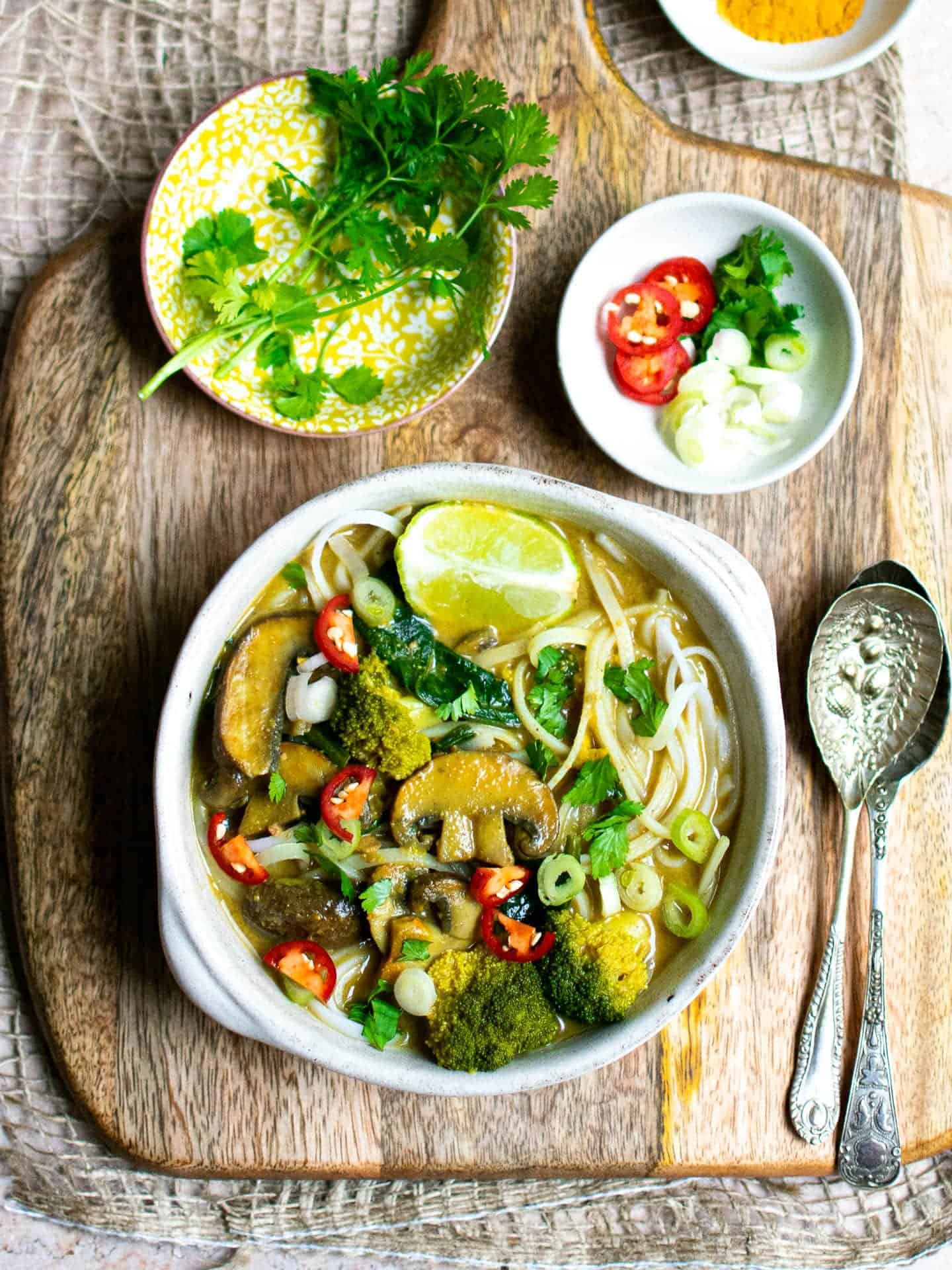 Noodle soup on a chopping board with spoons to the right, pinch bowls full of spring onions and fresh coriander