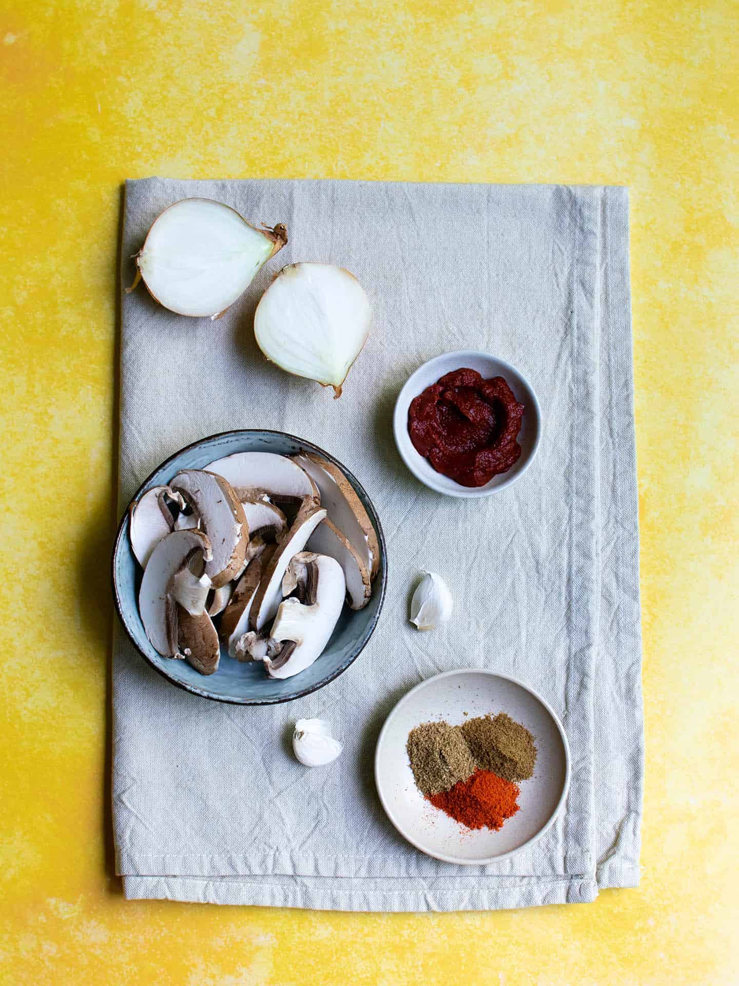 Ingredients for mushroom bhaji laid out on a white cloth, showing an onion, spices, mushrooms, garlic and spices