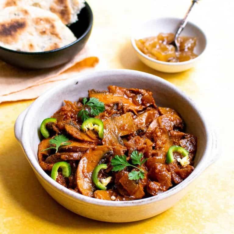 Mushroom bhaji in a small dish with chutney and naan bread in the background, all on a yellow background