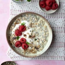 A bowl of overnight oats with raspberry, topped with yoghurt and granola. There's a small dish of raspberries and some mint behind the bowl