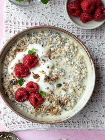 A bowl of overnight oats with raspberry, topped with yoghurt and granola. There's a small dish of raspberries and some mint behind the bowl