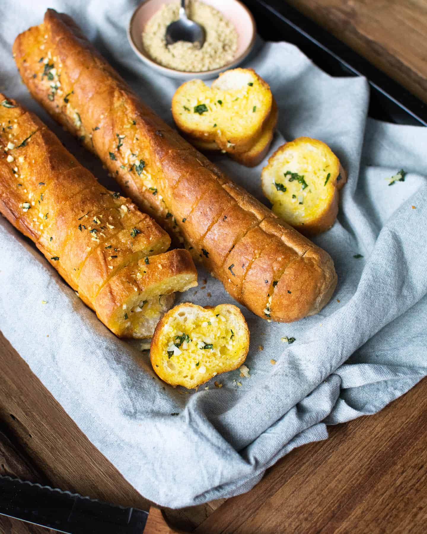 Top down view of 2 vegan garlic bread baguettes on a blue cloth, with three slices in the background with some cashew cheese in a pink dish