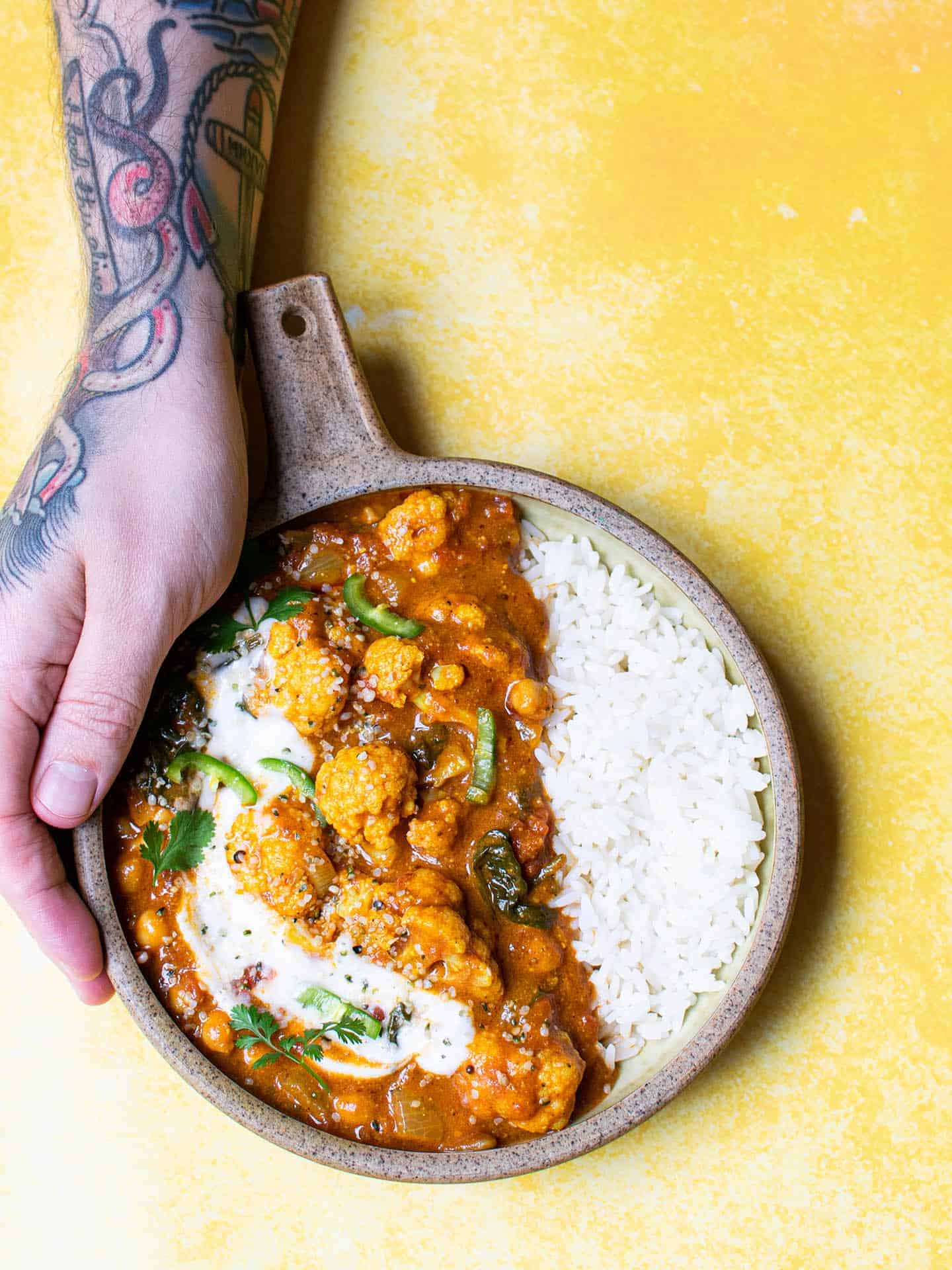 Vegan cauliflower curry in a bowl with a tattooed arm about to pick it up