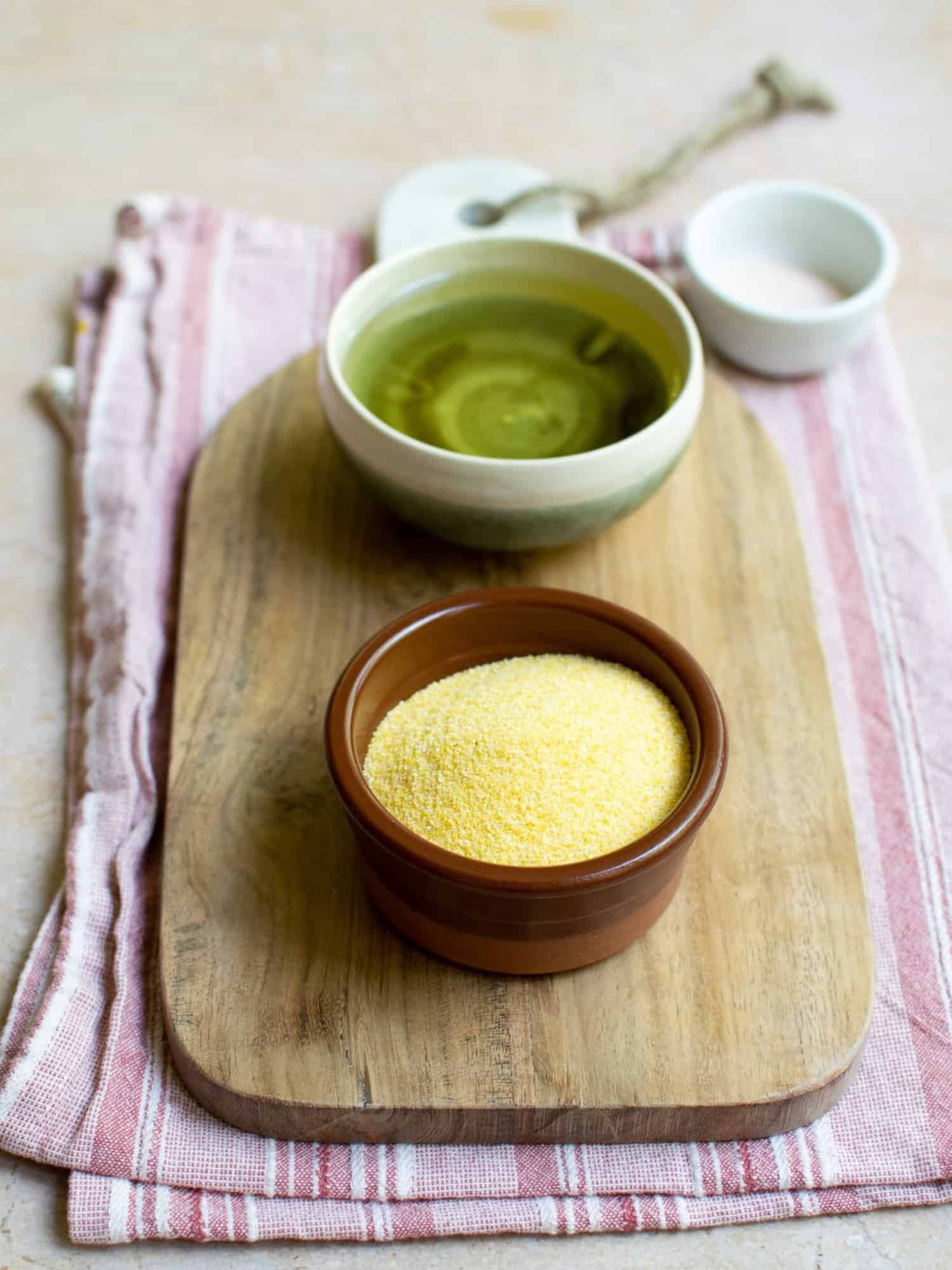 In two separate bowls there is polenta and oil. Set on a chopping board with a pink cloth underneath it