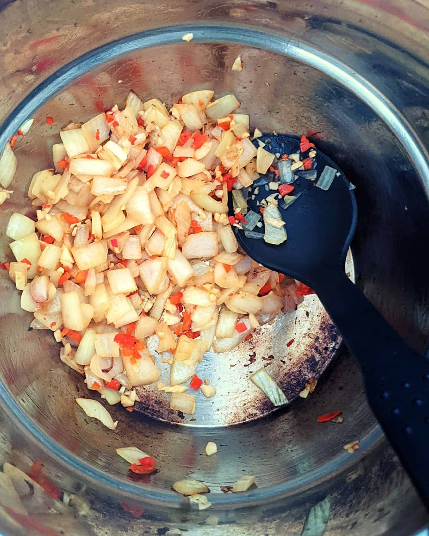 Chopped onions, chilli and garlic in a pan, with a black spoon resting in it