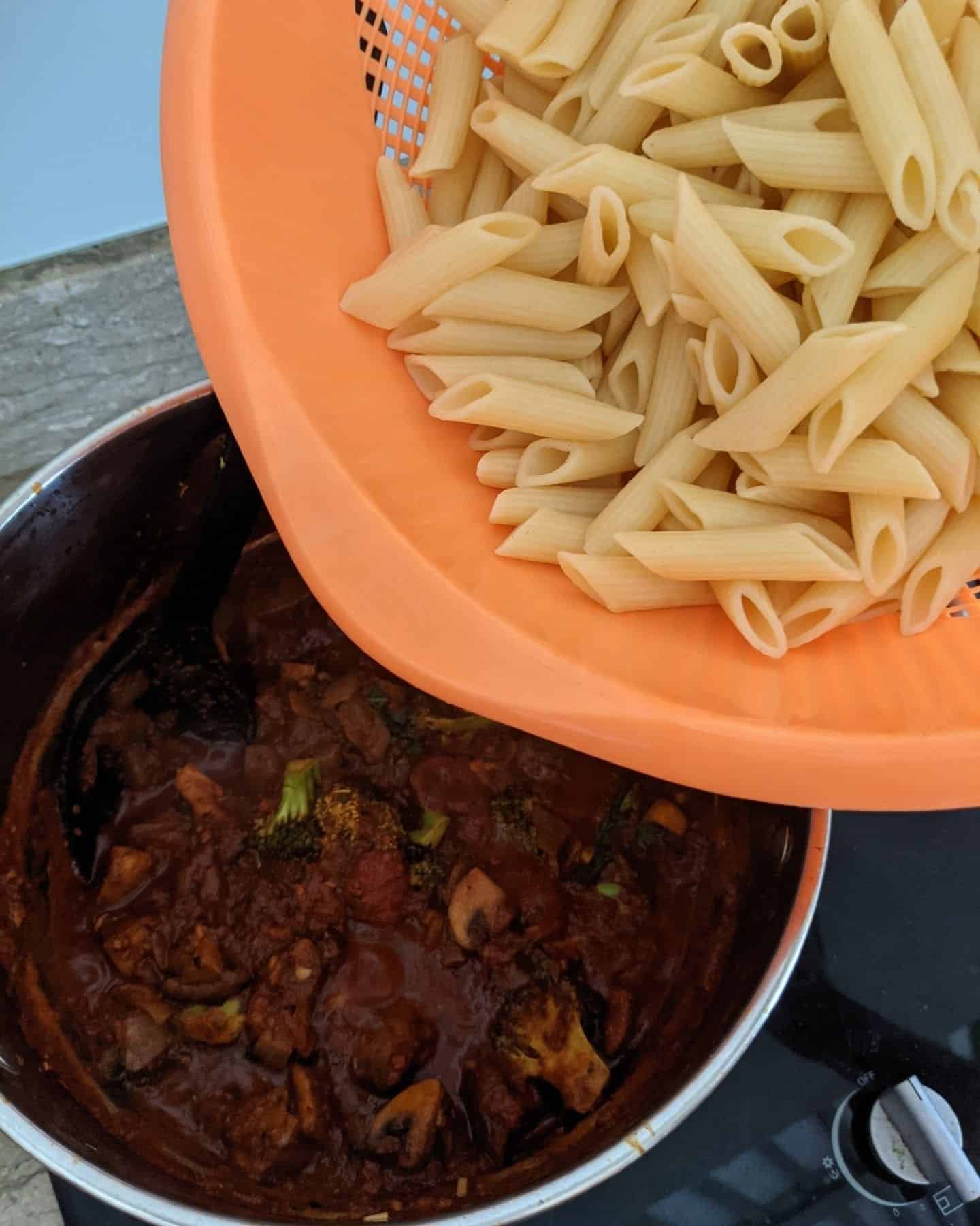 Penne pasta in an orange colander about to be poured into a pan of vegan arrabiata sauce