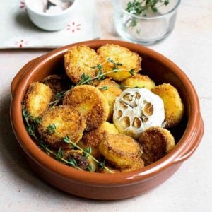 Roast potatoes in a ceramic dish with a whole roasted garlic and herbs in the background