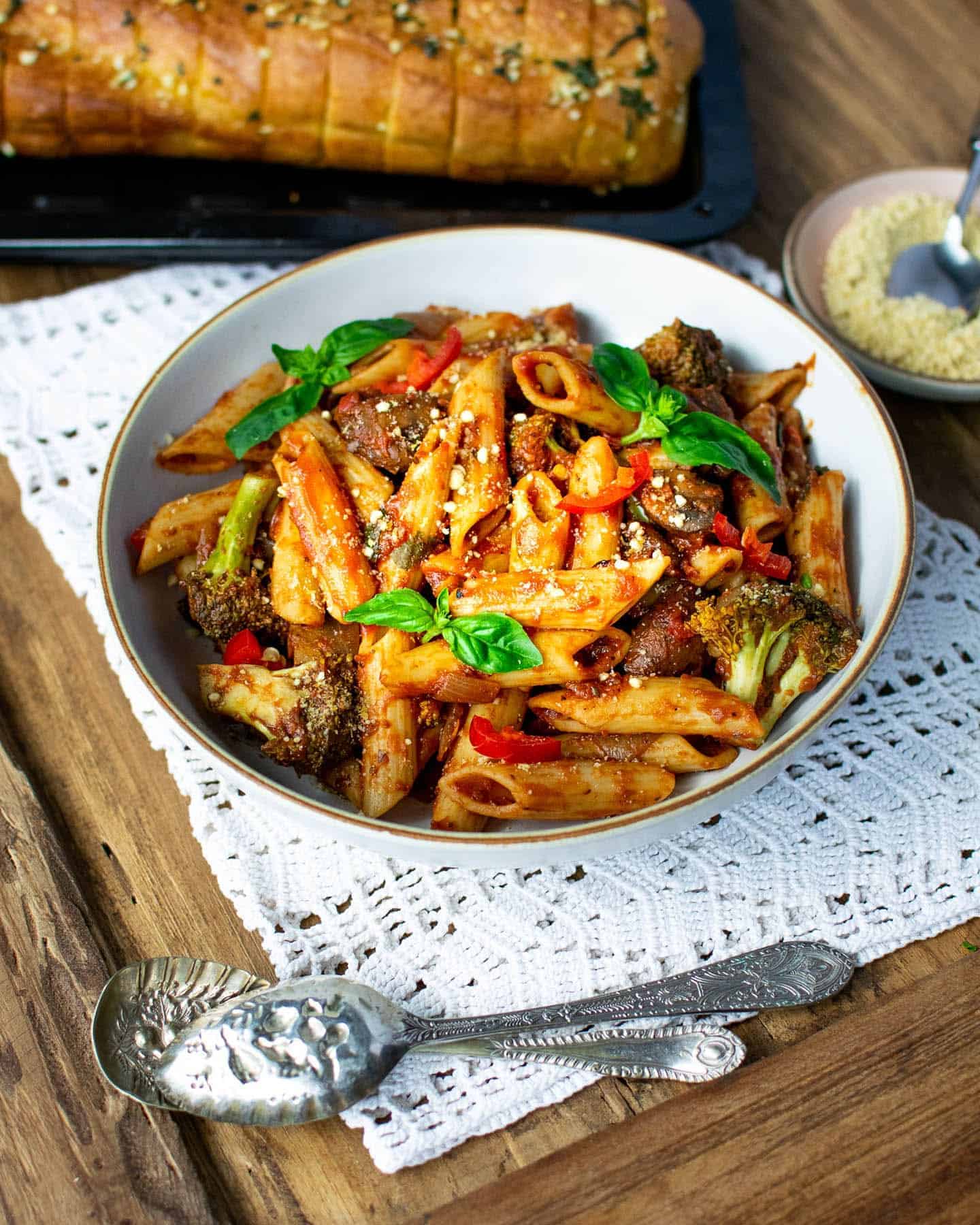 Diagonal view of a bowl of vegan arrabiata pasta with spoons in the foreground, fresh basil sprigs on top and garlic bread and nut parmesan in the background