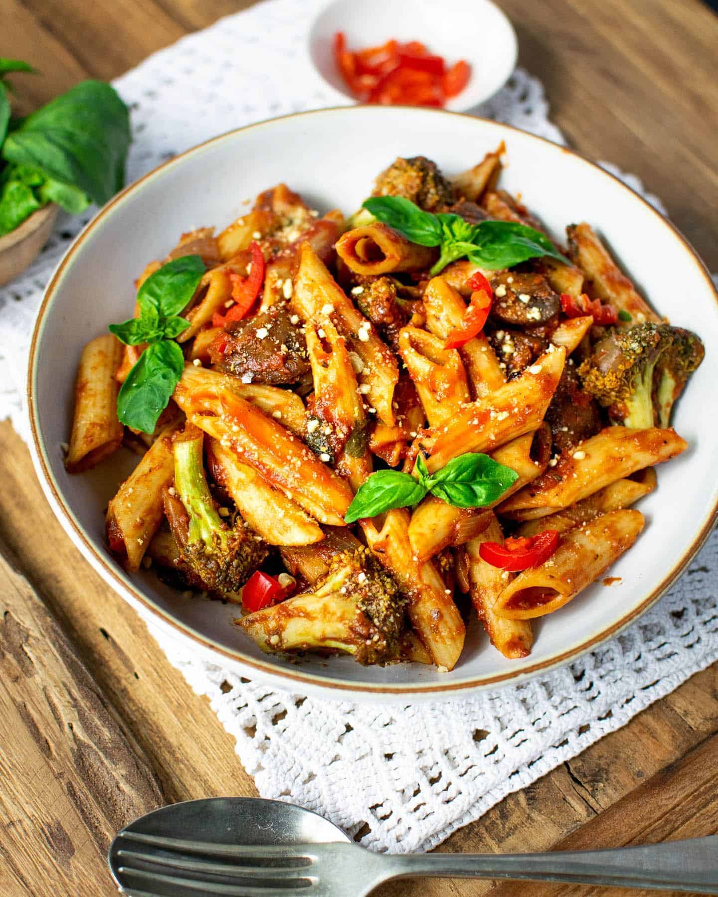 A white bowl of vegan arrabiata with fresh basil on top and a spoon and fork in the foreground with some fresh chilli in the background and some fresh basil in a wooden pot to the left hand side in the background