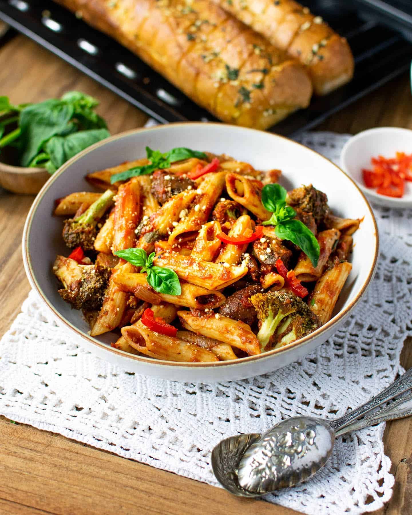 A bowl of pasta with garlic bread in the background, fresh red chilli in a small dish to the right and fresh basil to the left