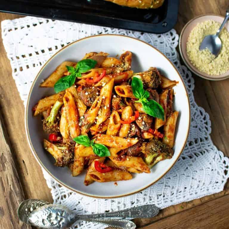 Top down view of vegan arrabiata pasta topped with basil, sitting on a white cloth on a wooden table, with nut parmesan to the right in the background and some gardic bread just behind the pasta