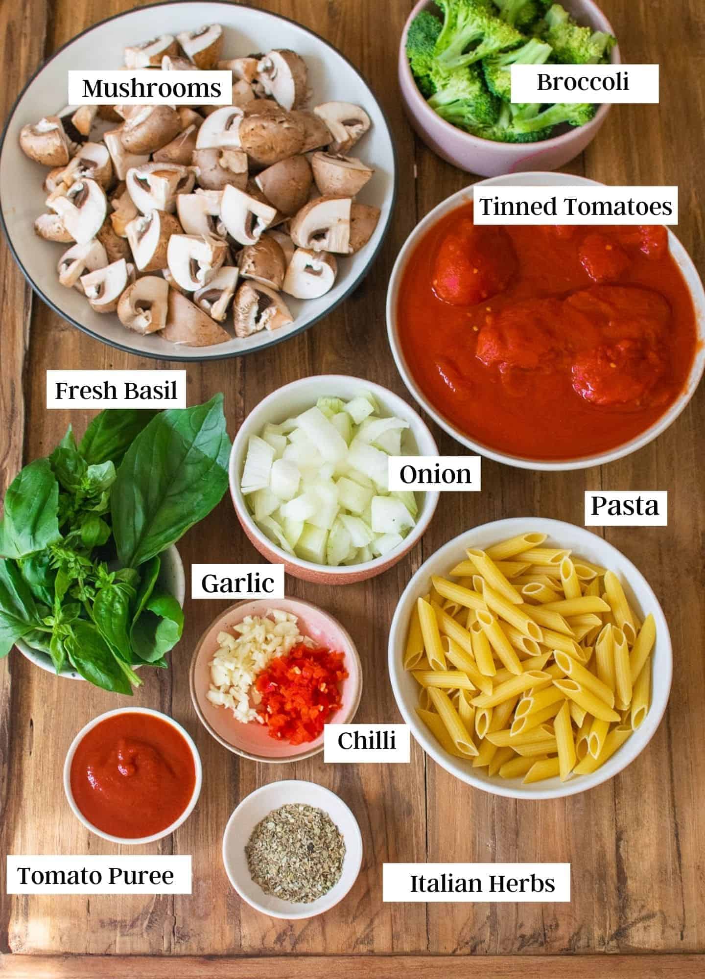 All of the ingredients for this recipe laid out on the table, and each item is labelled. It shows broccoli, tomatoes, mushrooms, onion, basil, chilli, garlic, pasta. tomato puree and Italian herbs