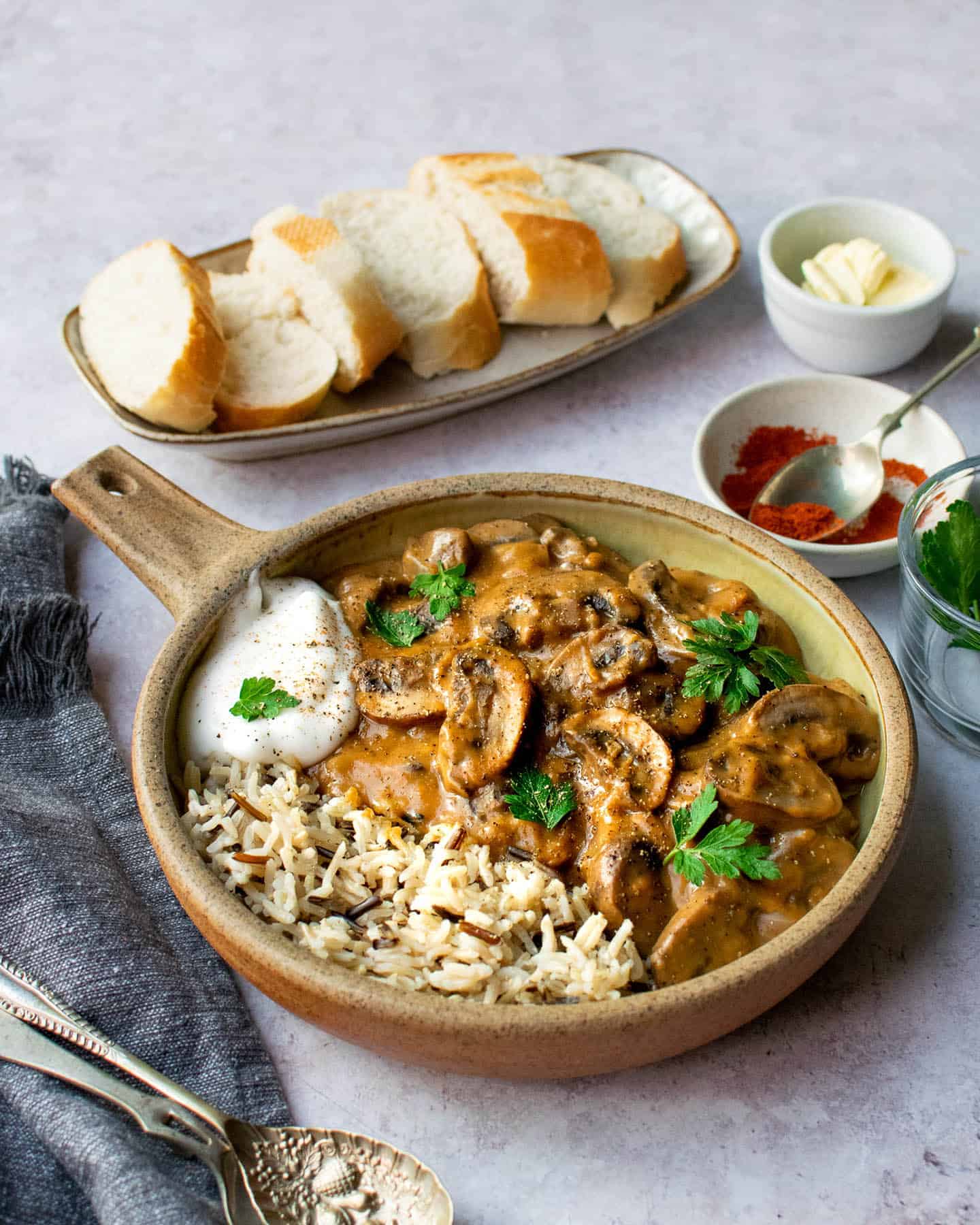 A bowl of wild rice and mushroom stroganoff. There's a plate of slices of crusty bread, as well as some bowls of spices and herbs surrounding it.