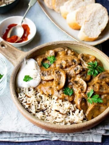 A bowl with a handle full with vegan stroganoff and wild rice with a dollop of vegan yoghurt on the side. Crusty bread behind it as well as parsley in a glass pot and paprika in a white pinch bowl