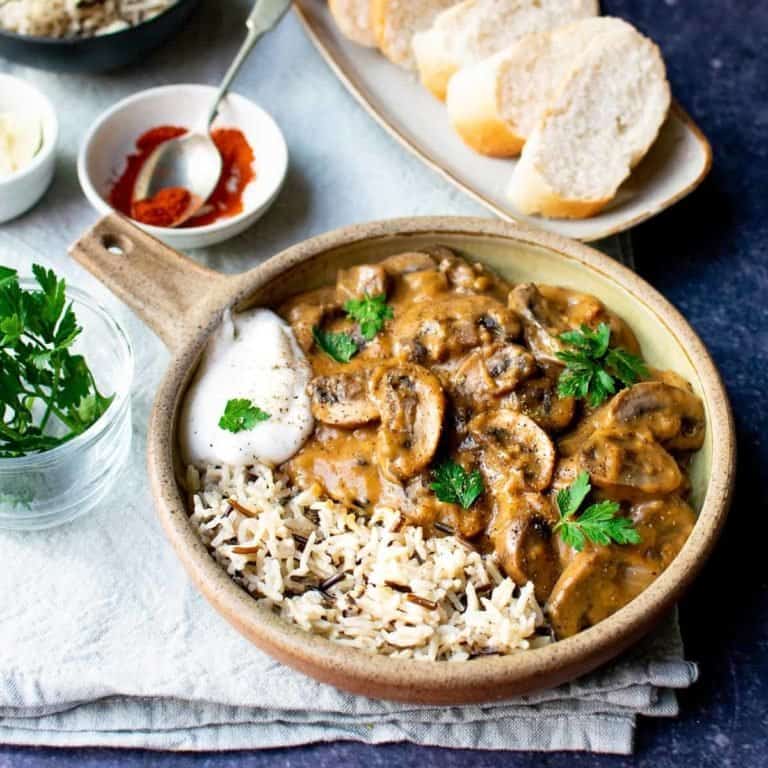 A bowl with a handle full with vegan stroganoff and wild rice with a dollop of vegan yoghurt on the side. Crusty bread behind it as well as parsley in a glass pot and paprika in a white pinch bowl