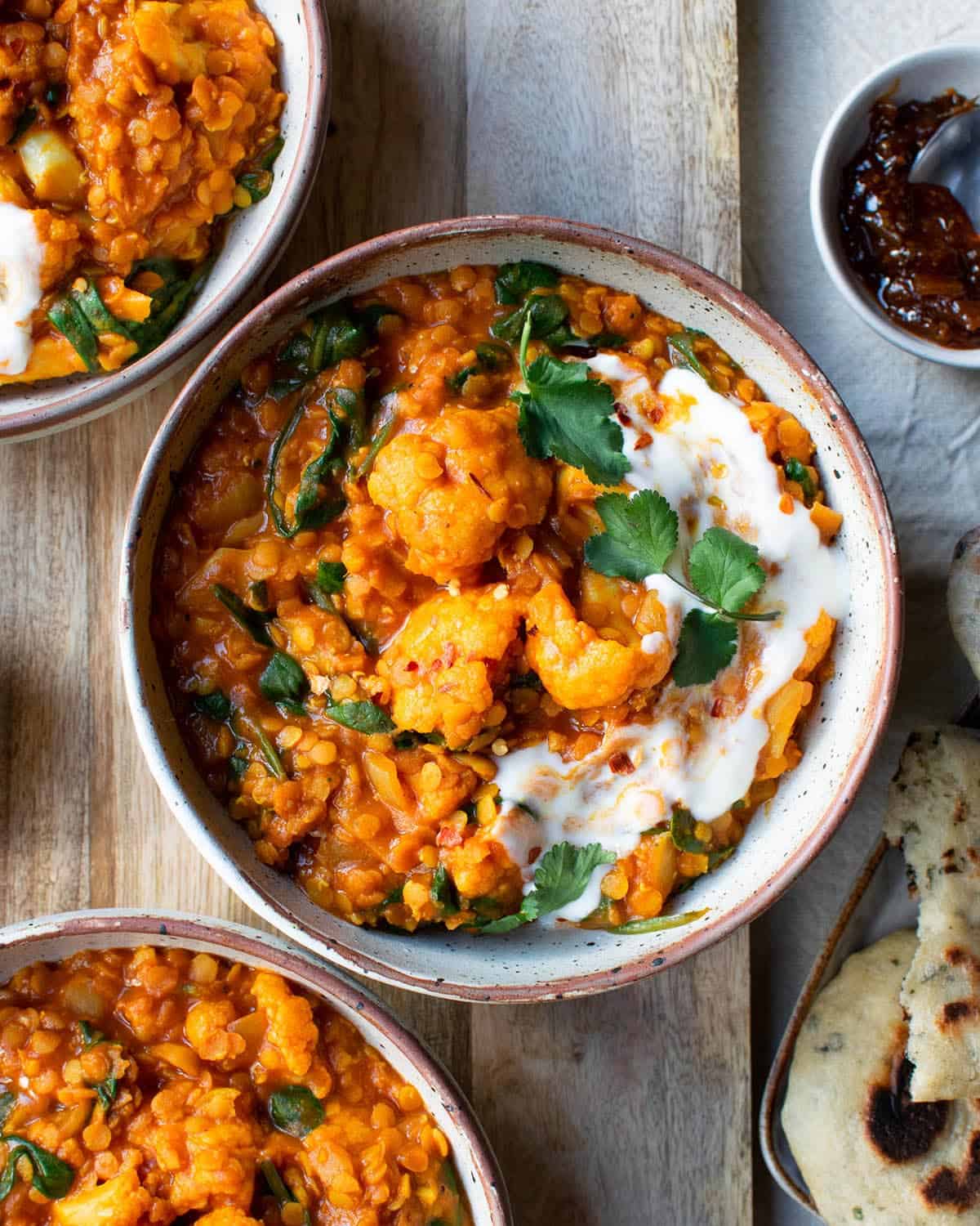 Red lentil curry in a bowl with two more bowls of curry, chutney and naan.