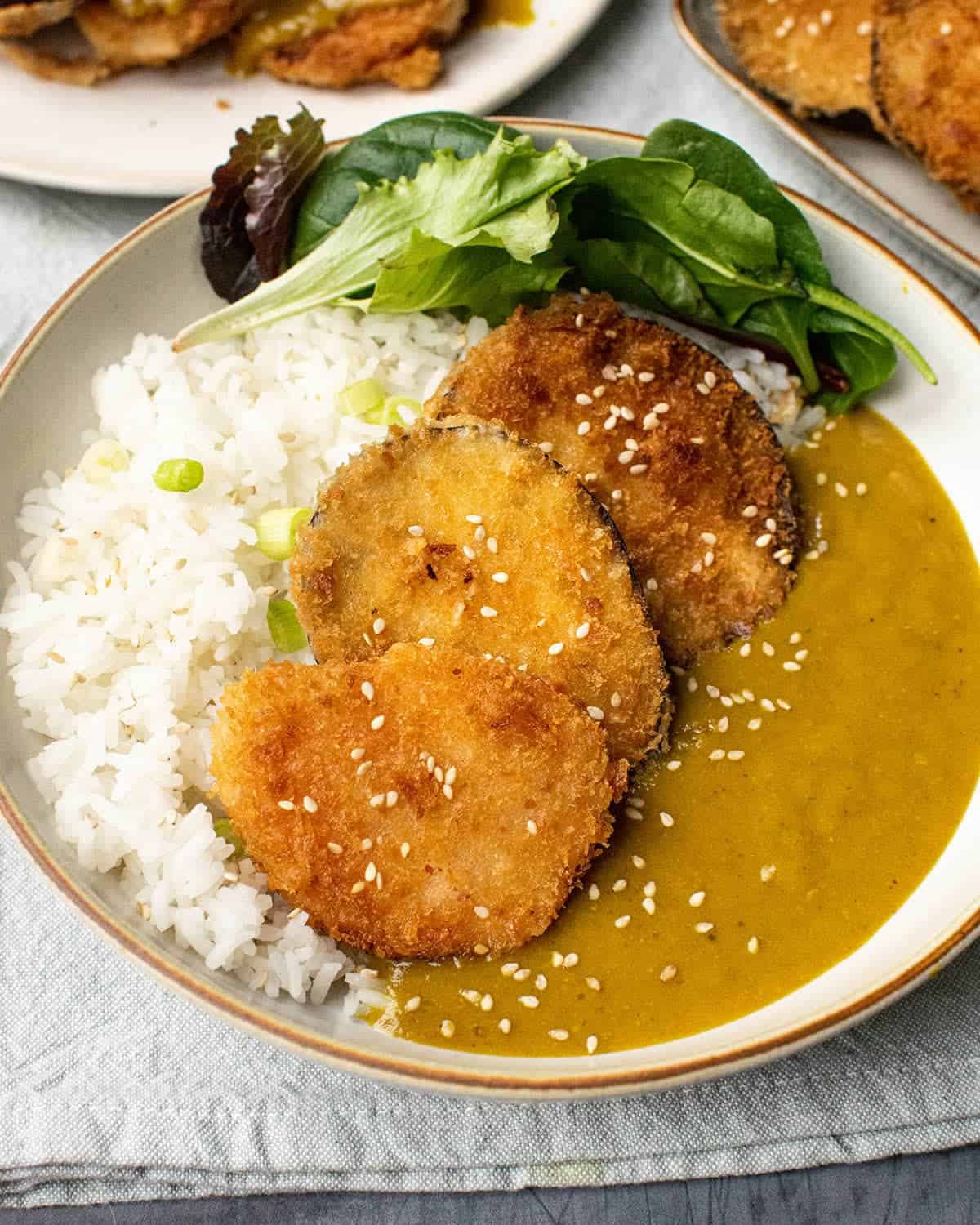 Close up of aubergine and sweet potato katsu curry served with curry sauce, salad and white rice.
