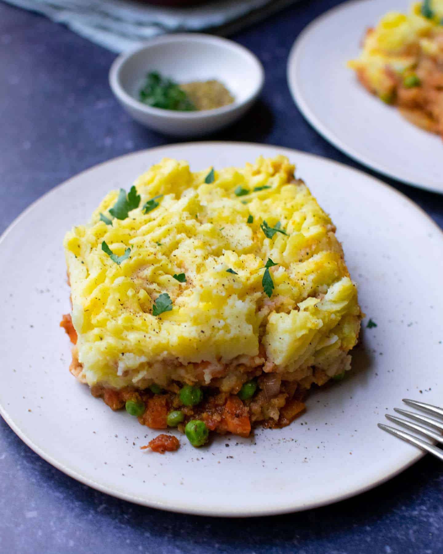 A close up of TVP shepherd's pie on a white plate with a pinch bowl in the background. There's parsley on top of the mashed potato (which is the topping)