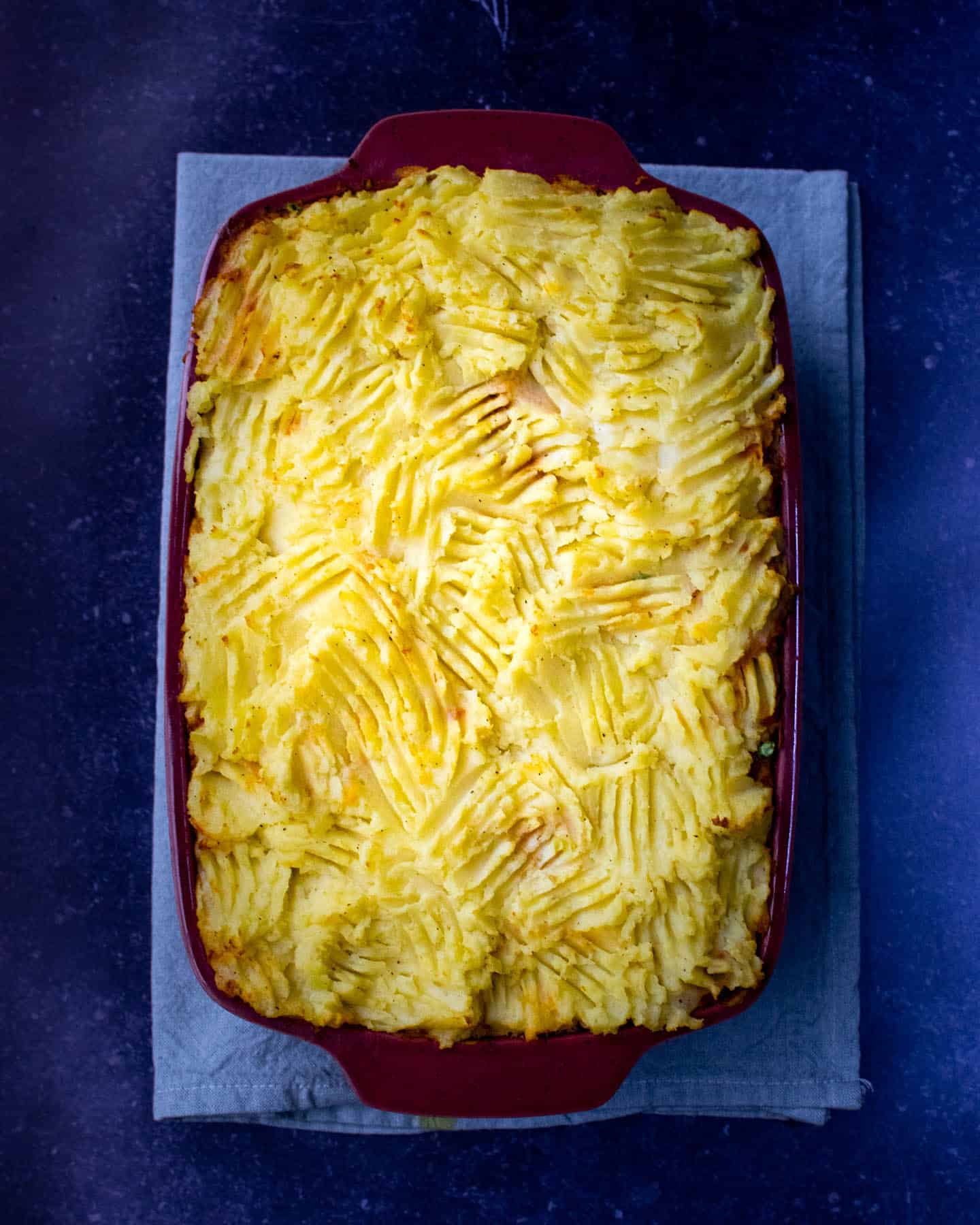 A top down view of a red baking dish full to the top with mash potato that has fork imprints all over it. All set on a dark blue background