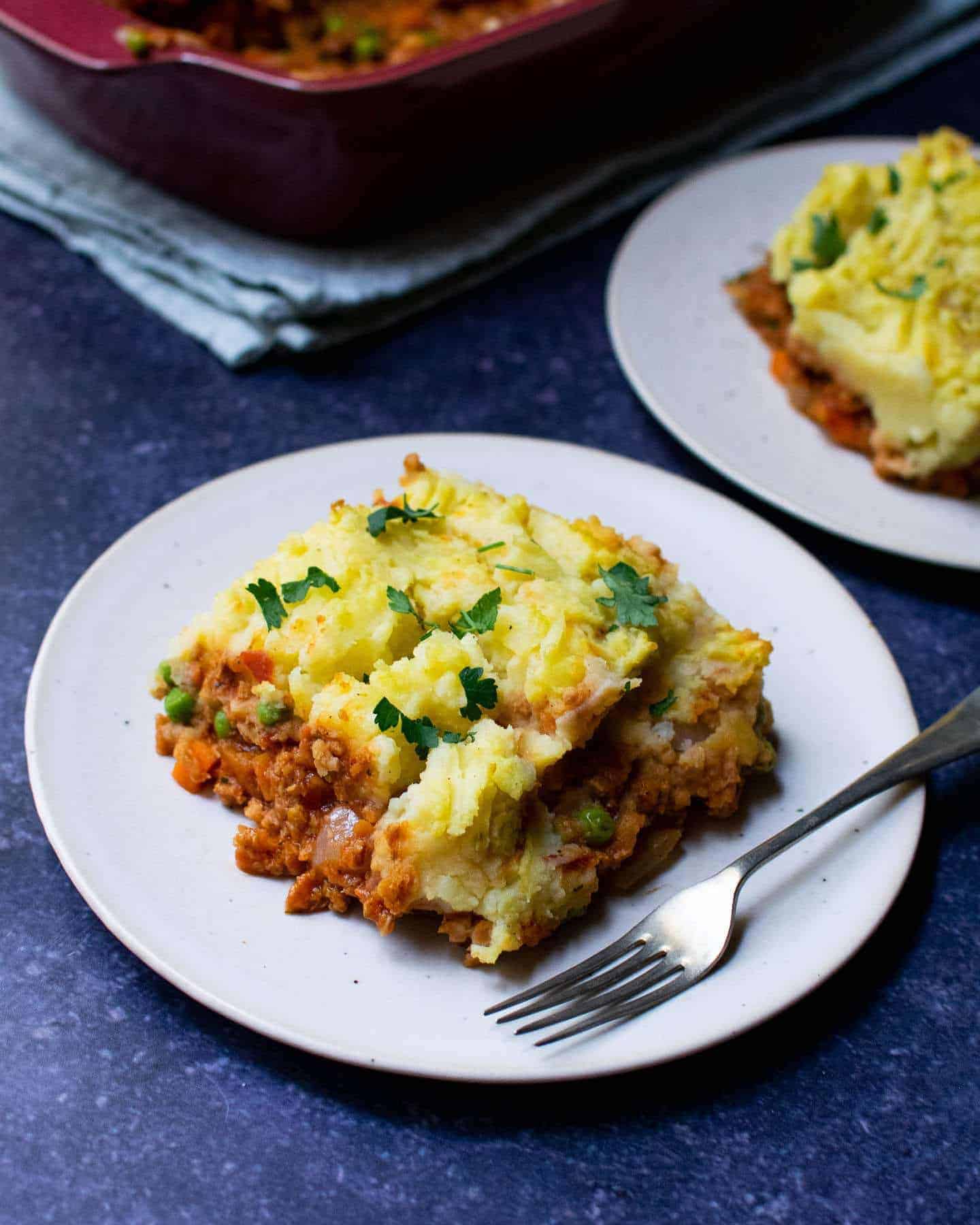 A slice of TVP shepherd's pie on a white plate, topped with mashed potatoes and parsley. There's a fork resting on the plate and another plate and a dish in the background