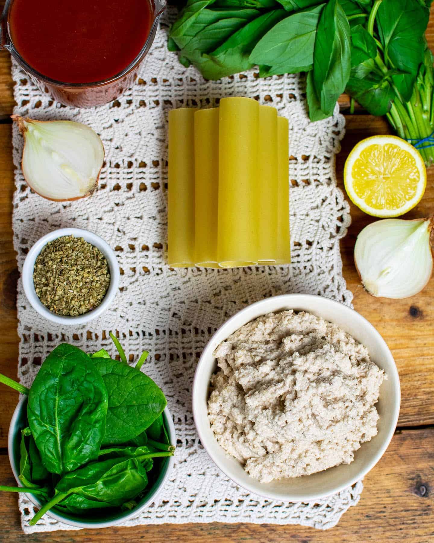 Top down view of ingredients laid out on white fabric, showing pasta tubes, macadamia ricotta, onion, lemon, spinach, fresh basil, dried oregano and passata