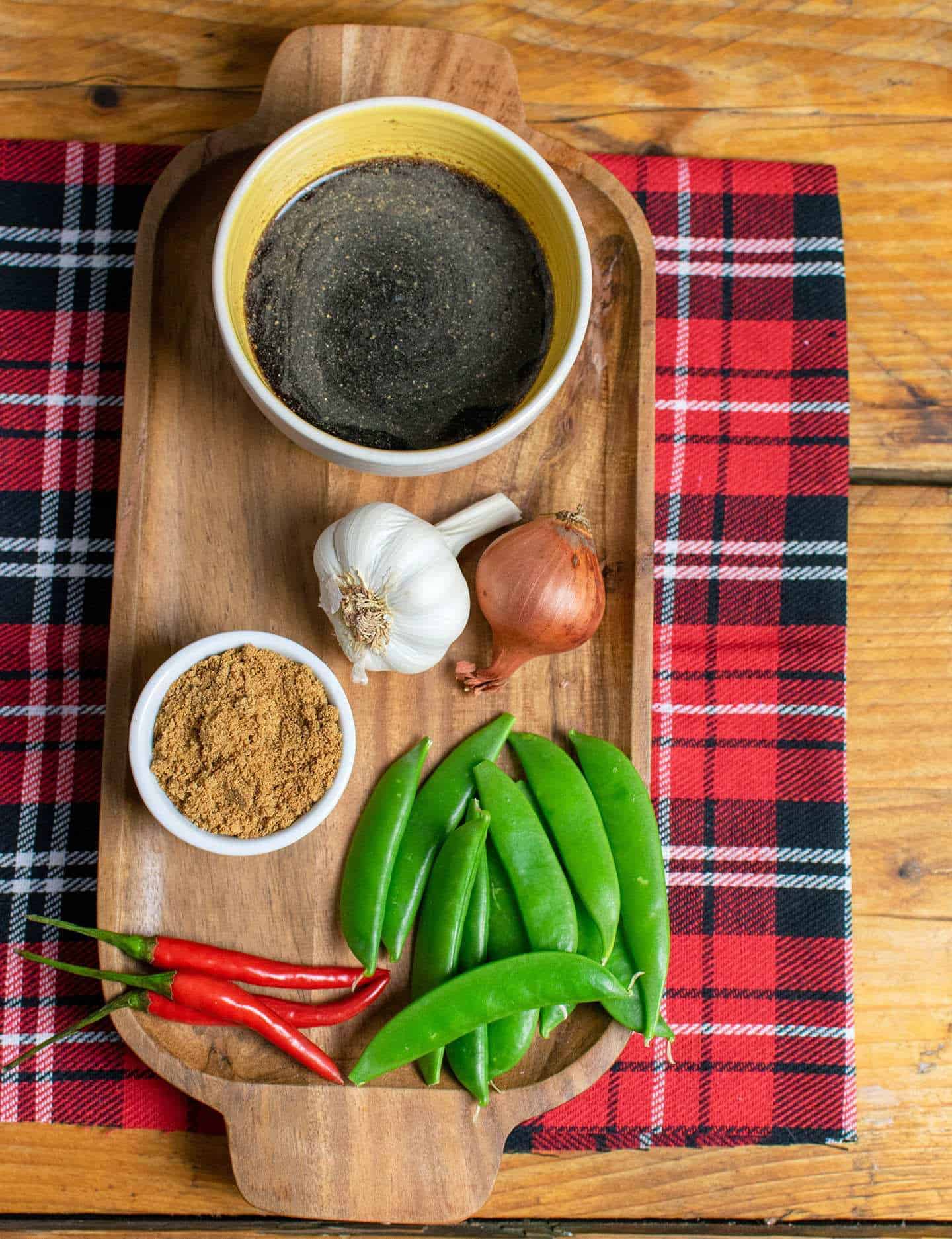 Ingredients laid out on a wooden try, including sauce, sugar, garlic, onion, sugar snap peas and chillies