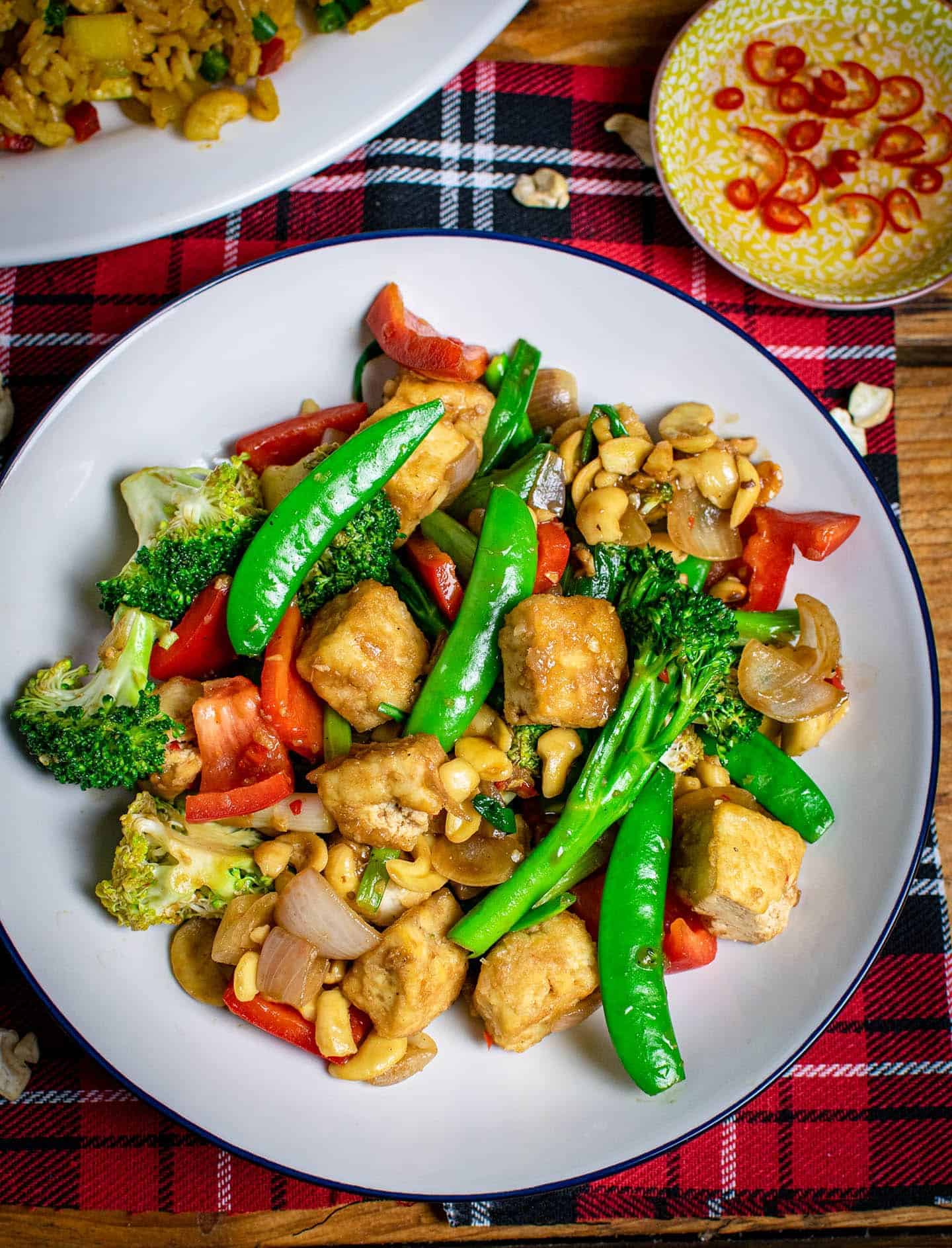 Thai cashew stir fry on a white plate with chillies on a small yellow bowl in the background, and rice in the top left corner.