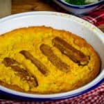 Vegan Toad in the Hole in a white enamel oven dish with a blue rim. Knife and fork to the right of the dish, a read and white gingham cloth surrounding it and assorted vegetables in a dish in the right hand corner of the image.