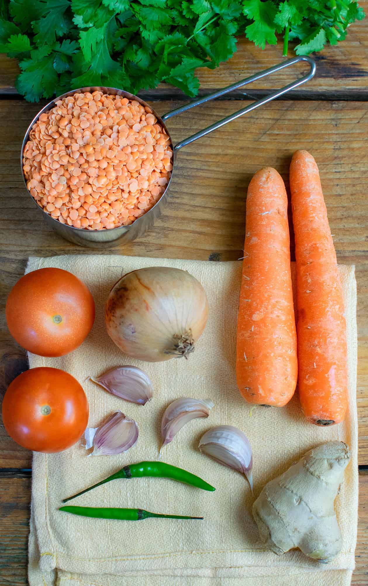 Ingredients laid out on an orange cloth, showing coriander, carrots, red split lentils, onion, tomatoes, garlic, ginger and green chillies.