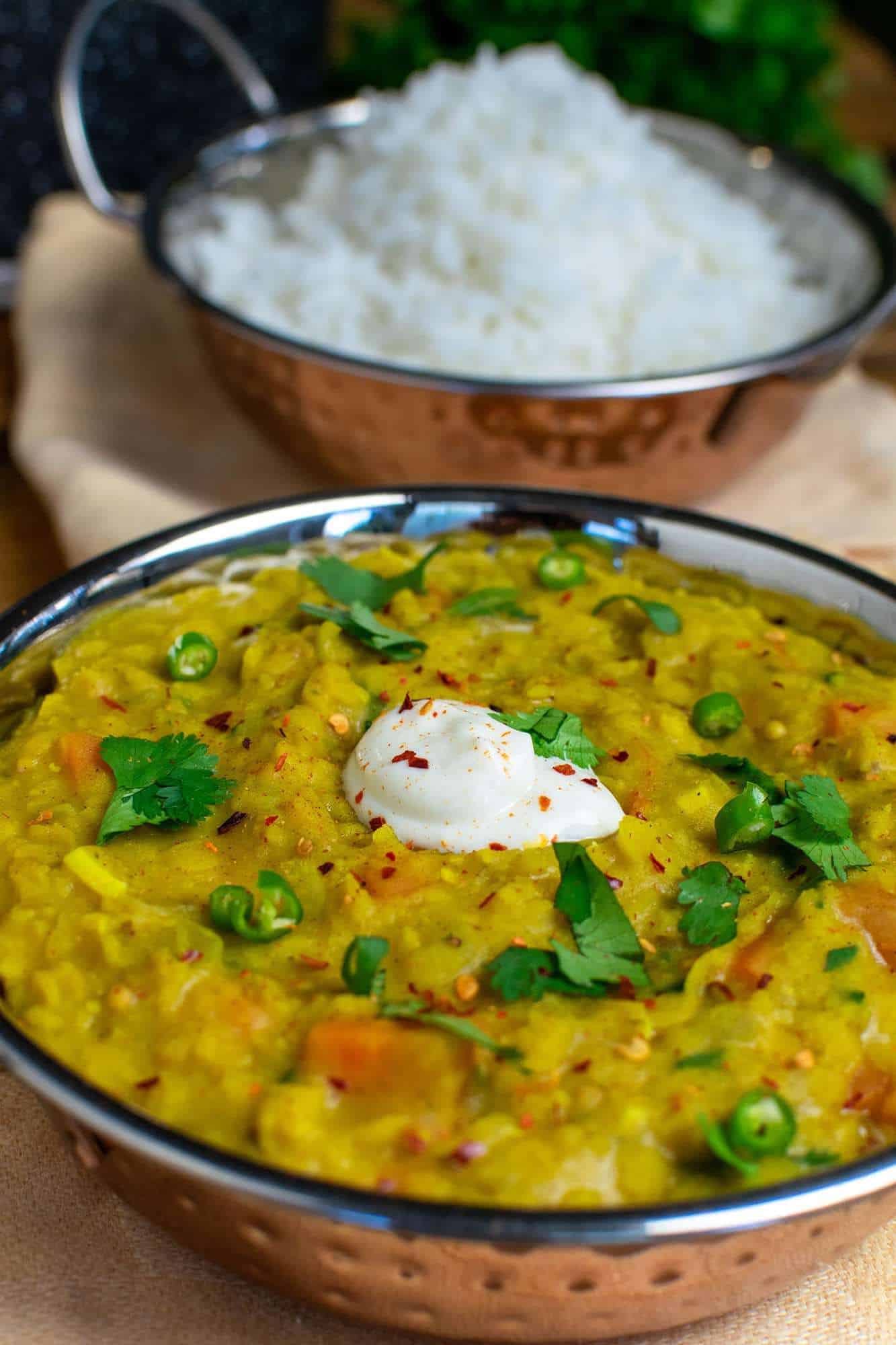 Vegan dahl piled high in an Indian metalic dish with white rice in the background and a pan is just visible. 