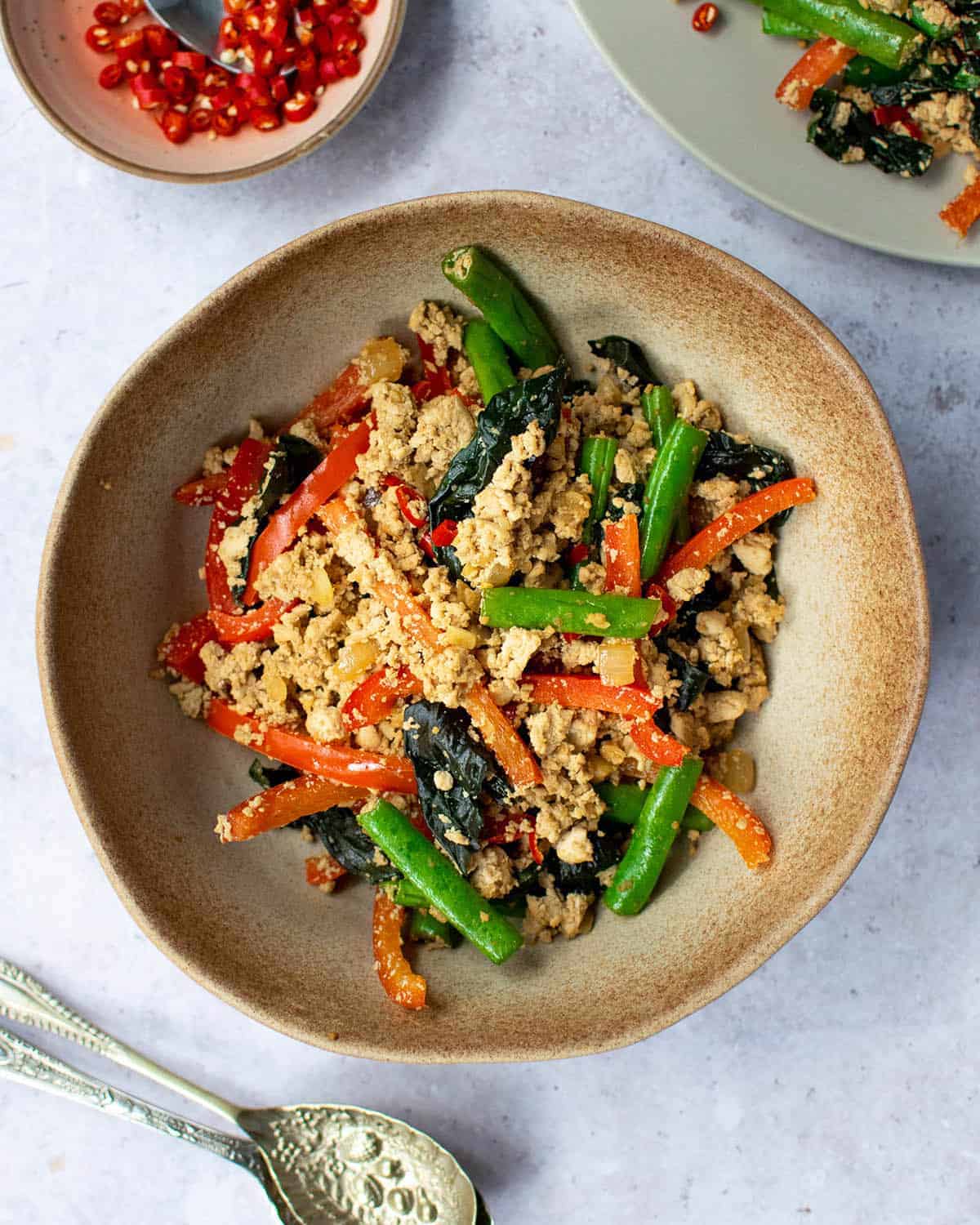 Top down view of tofu basil stir fry in a brown bowl with chillies in a pot above it