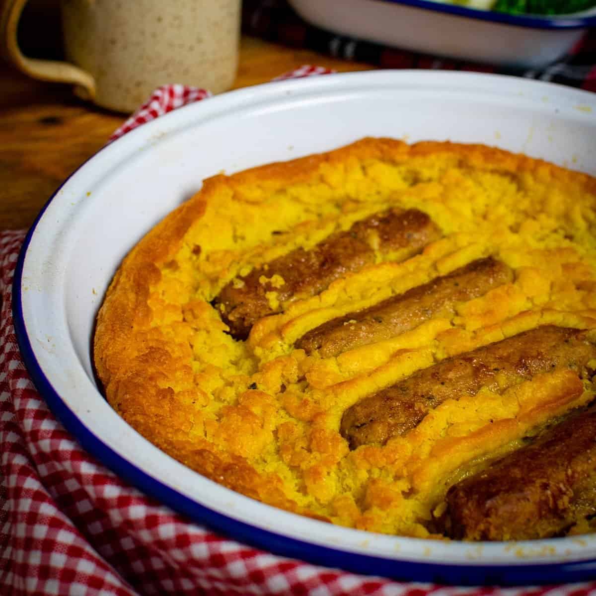 Vegan Toad in the Hole in a white and blue round oven dish, showing 4 vegan sausages and perfecty golden batter surrounding the sausages. There's a red and white gingham cloth surrounding the oven dish.