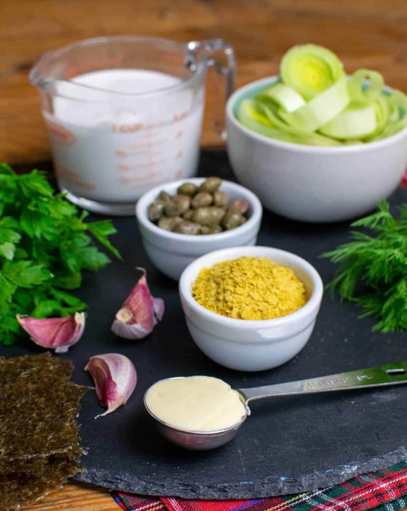 Ingredients laid out on a round slate. Including nutritional yeast, capers, vegan butter, garlic, leeks, parsley and dill.
