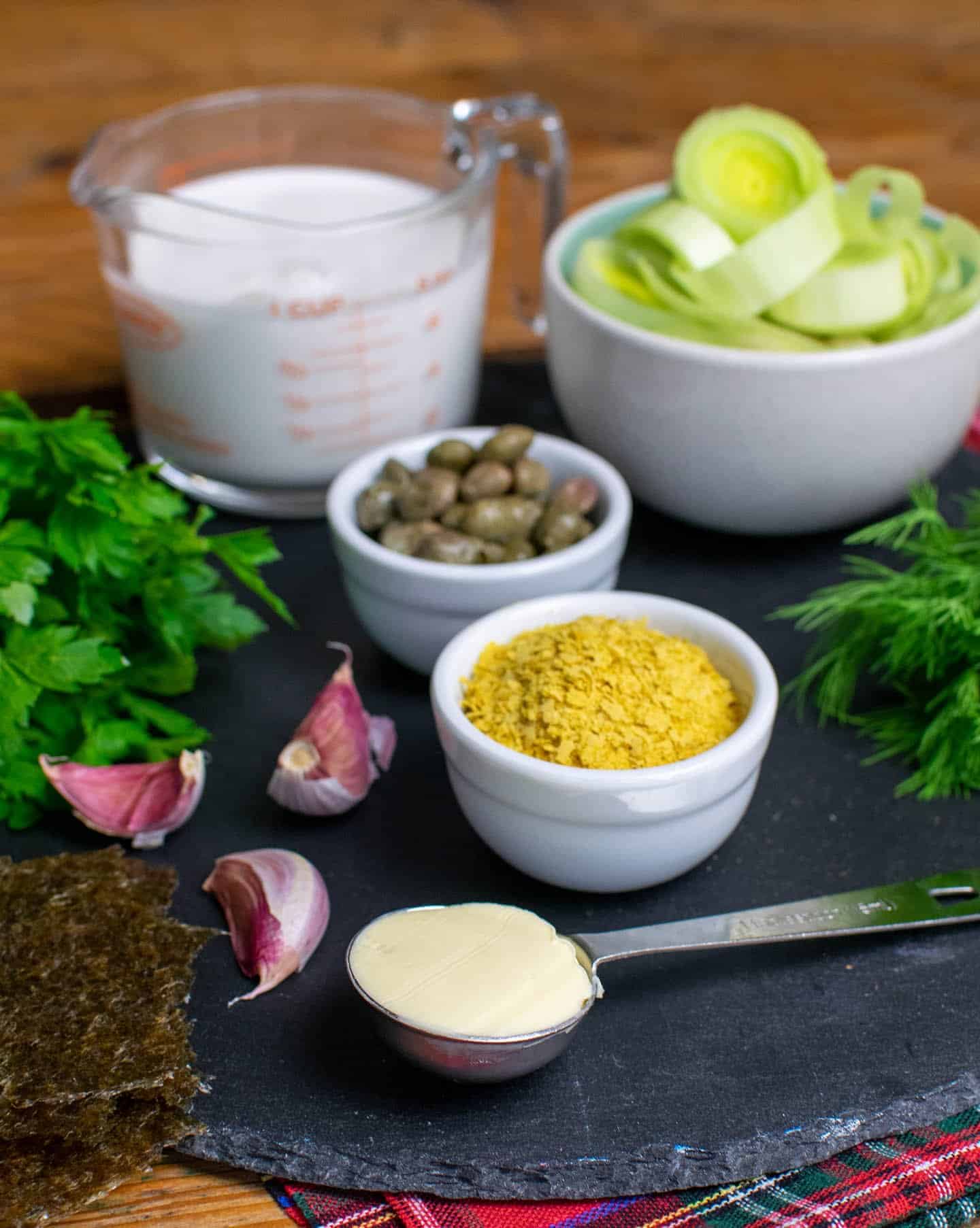 A round slate board with small pots of nutritional yeast, capers and leeks. A tablespoon is full of vegan butter, cloves of garlic, parsley and dill also visible. 