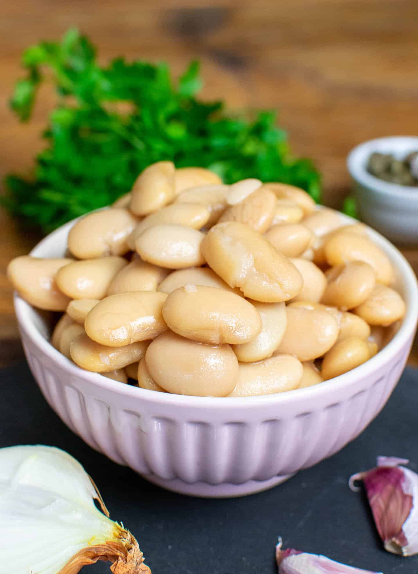 A pink bowl of butter beans piled high, a bunch pf parley is in the background with garlic and onion in the foreground.