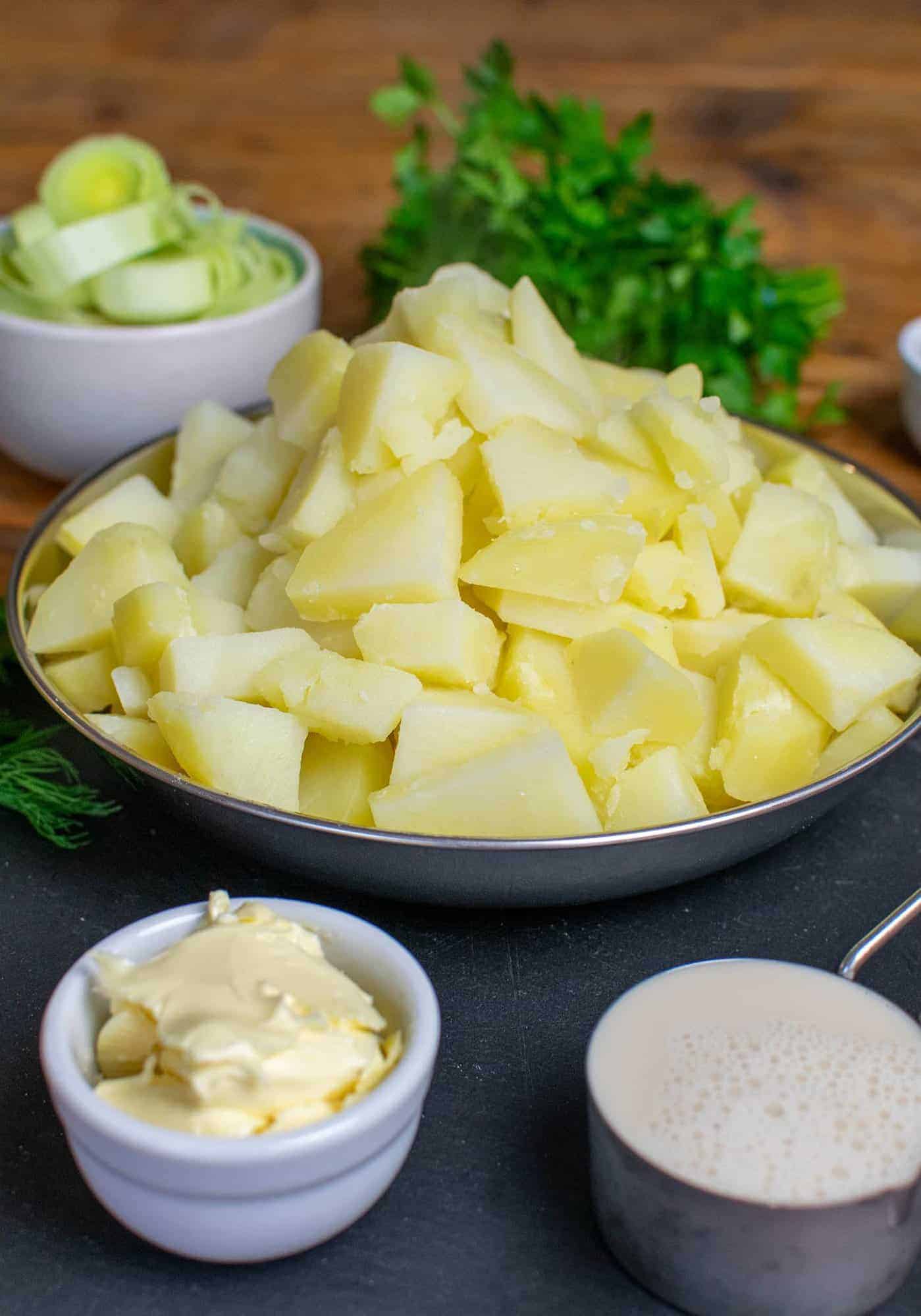 A big pile of cooked potatoes on a silver tray with oat milk in a quarter cup measurer, vegan butter in a white pot and leeks and parsley in the background