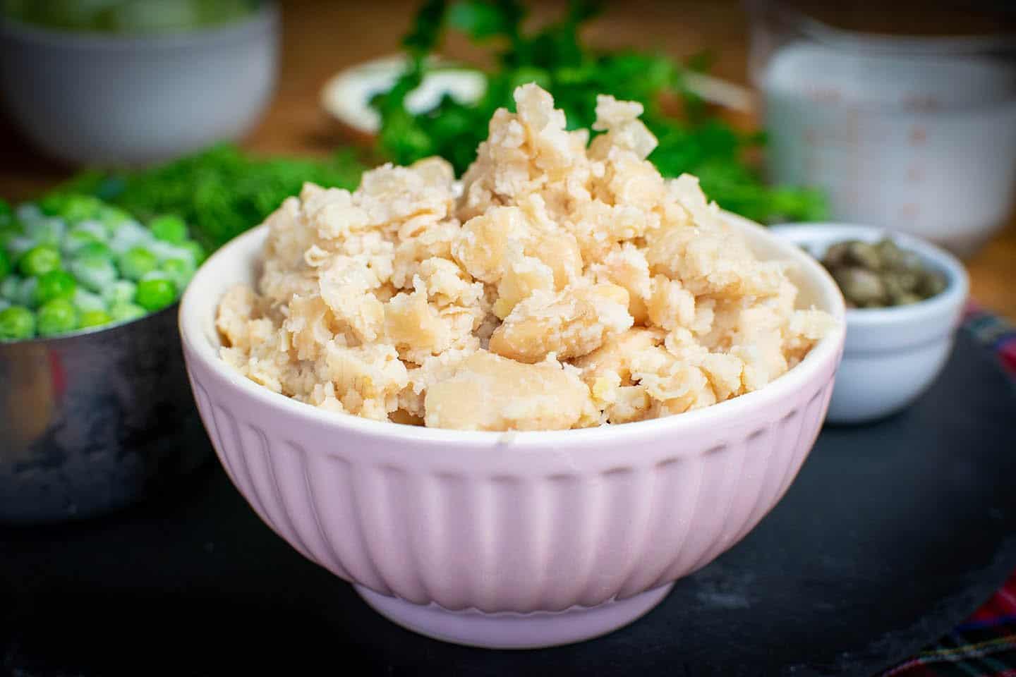 A pink bowl full of mashed butter beans with parsley, peas and capers in the background.