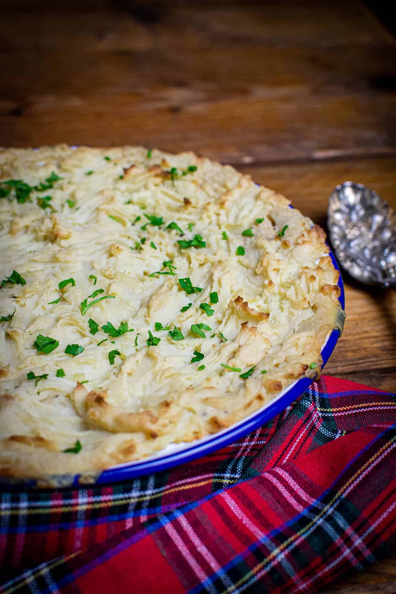 Fishless pie in a white round dish with a blue rim. There's a spoon in the background and a checked cloth in the foreground around the circular dish