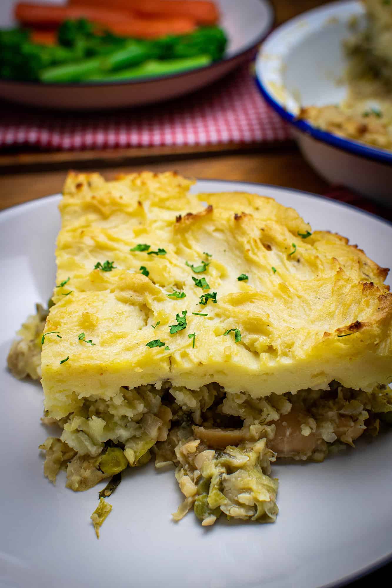 A piece of fishless pie on a cream plate with parsley on top of the mashed potato. The pie dish and vegetables can be seen in the background.