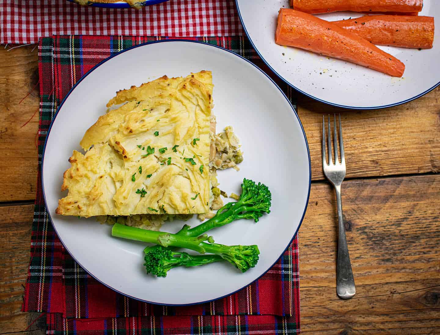 A slice of vegan fish pie with tenderstem broccoli on a cream plate with a fork to the right. Carrots in the top right corner on another cream plate.